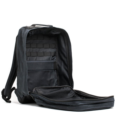 The GR2 - Waxed Canvas by GORUCK is open, displaying its spacious interior with multiple compartments and pockets. The black backpack is propped upright, highlighting its sturdy design and padded shoulder straps. Inside, it is neatly organized with elastic loops and mesh sections.