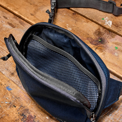 The compact black Belt Bag, crafted from durable Cordura and featuring a convenient mesh pocket, rests elegantly on a wooden surface.