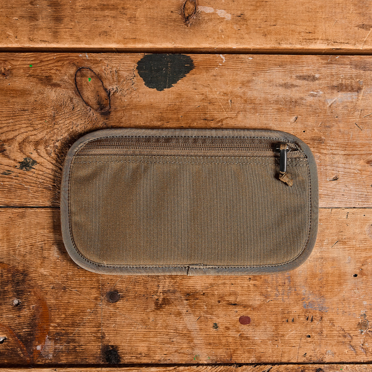 A GORUCK Admin Panels pouch, crafted from khaki fabric with a durable zipper, is ideal for travel or everyday carry essentials. It sits on a rustic wooden surface. The rectangular design is accented by visible stitching along its edges, while the wood beneath displays a textured look with assorted scratches and paint marks.