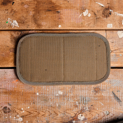 A GORUCK Admin Panel in a rectangular design with a textured beige surface lies on a wooden platform speckled with visible paint stains. The panel features rounded edges and two vertical stitching lines, while the wood displays its natural grain pattern accented with paint splatters.