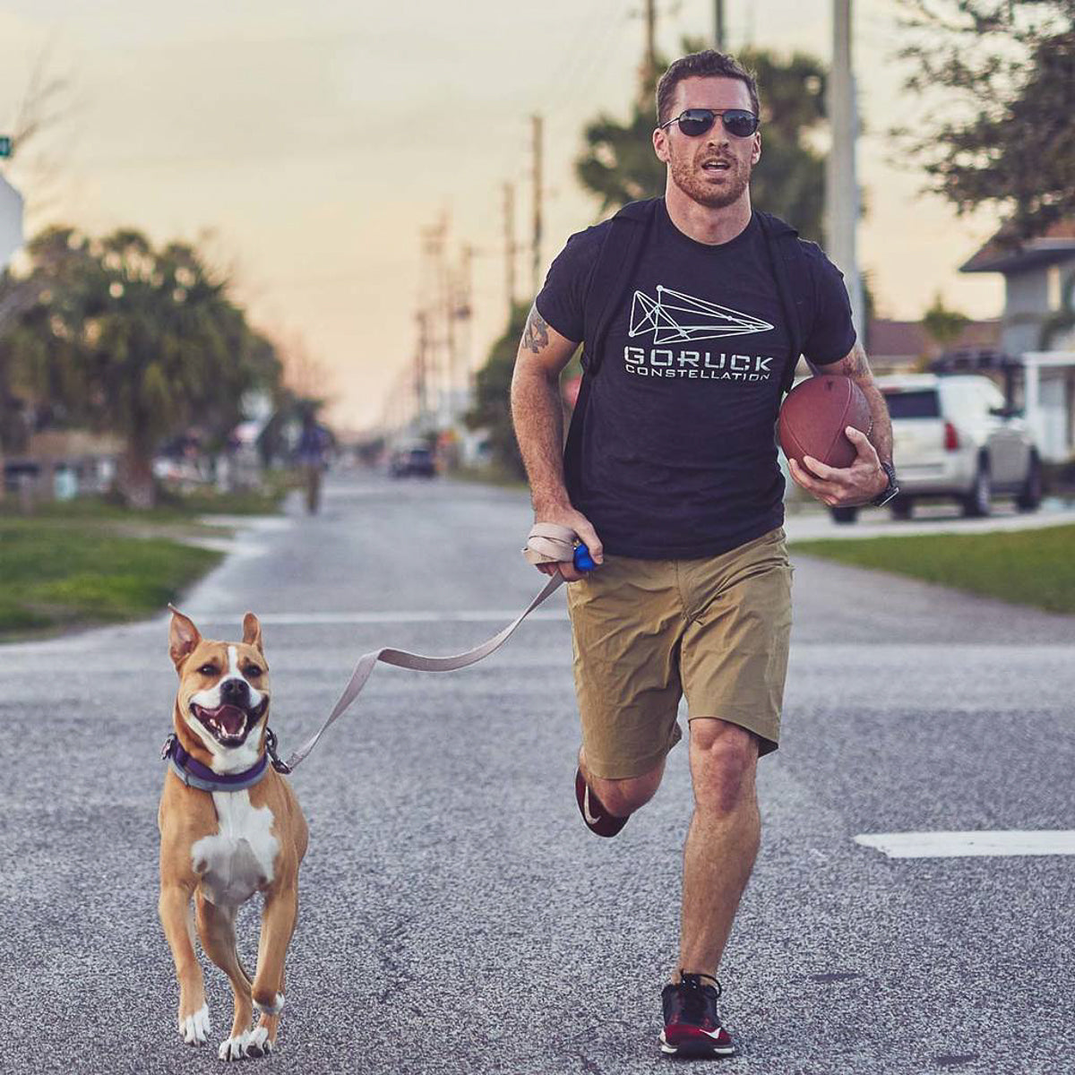 A man runs down a suburban street at sunset, clutching a football with his dog on a leash. Wearing Men’s Simple Shorts - Lightweight ToughDry®, he moves effortlessly, prepared for any adventure the glowing twilight may bring.