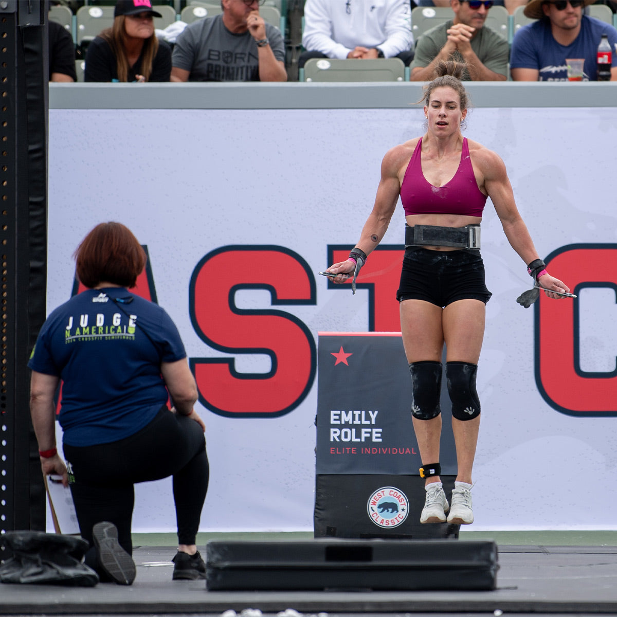 Dressed in a maroon sports bra and black shorts, a female athlete performs a jump rope exercise wearing GORUCK’s Women's Ballistic Trainers in Lunar Rock + Gum with Silver Reflective Spearhead. A judge kneels nearby, intently observing her agile performance as the name sign and audience fill the backdrop.