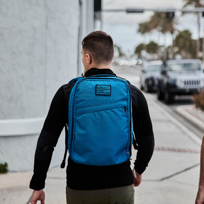 A person walks down a street wearing a black jacket and carrying an ultra lightweight, water-resistant blue backpack made from GORUCK's Ripstop ROBIC® material.