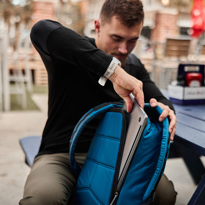 A person is placing an ultra-lightweight laptop into a blue, water-resistant GORUCK GR1 USA - Ripstop ROBIC® backpack while sitting at an outdoor table.