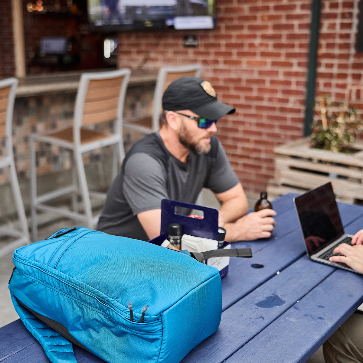 A man wearing a cap and sunglasses sits at a table with a GR1 USA - Ripstop ROBIC® bag from GORUCK, along with a laptop, holding a bottle.