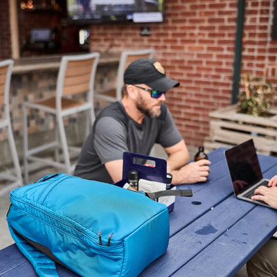 A man sits at an outdoor table with drinks, while his GR1 USA - Ripstop ROBIC® ultra lightweight blue backpack from GORUCK, made from water-resistant 420D Robic® Ripstop Nylon, rests in the foreground.