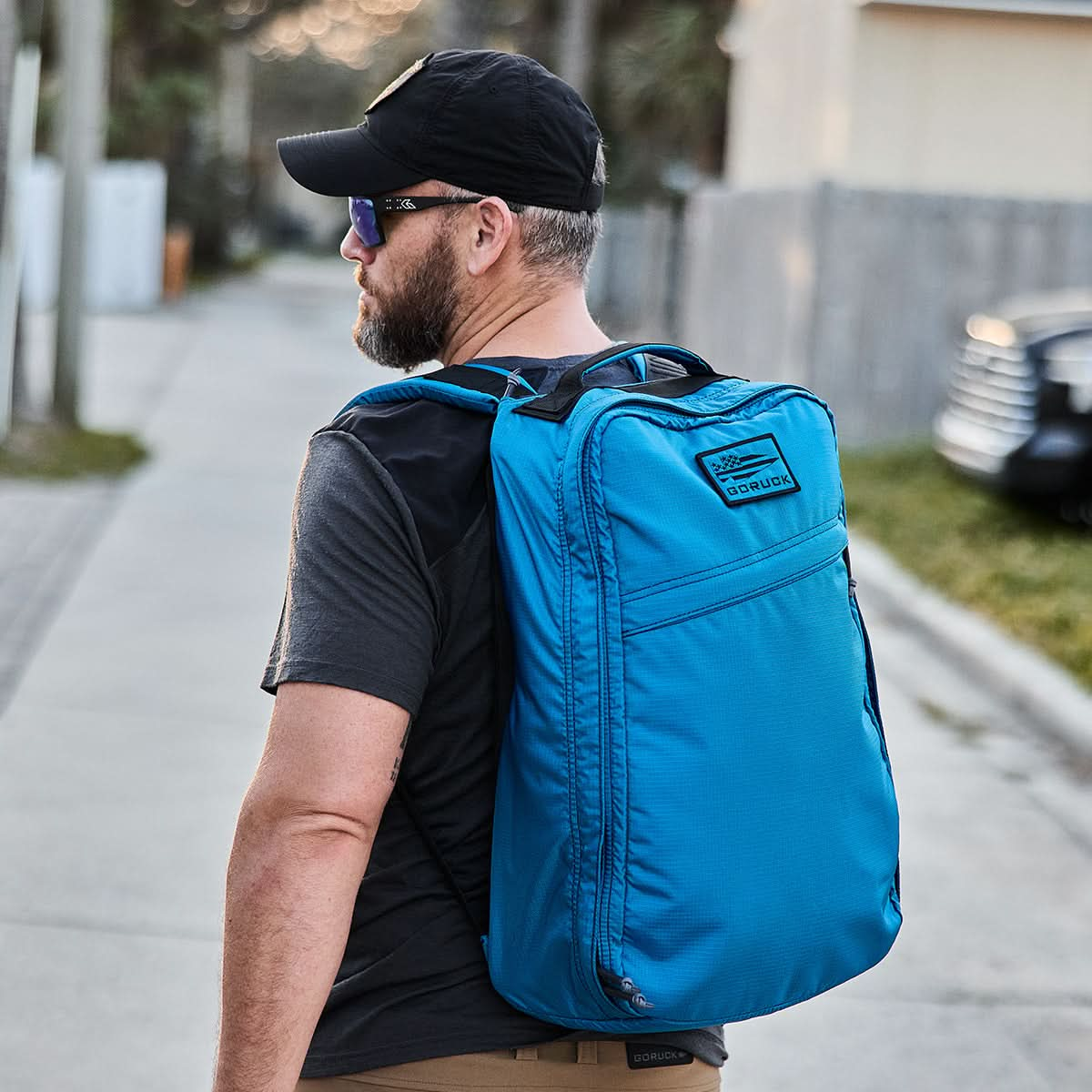 A man wearing sunglasses and a black cap strolls down a residential street, effortlessly carrying a GR1 USA - Ripstop ROBIC® from GORUCK. The large blue bag, made from water-resistant 420D Robic® Ripstop Nylon, is perfect for his urban adventure.