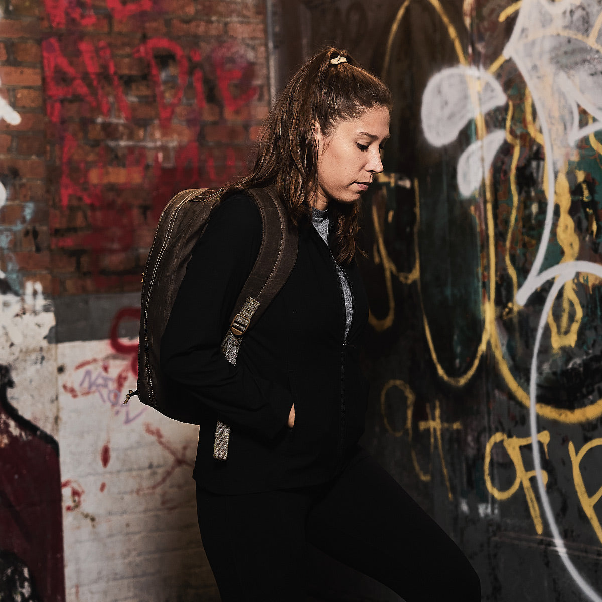 A woman donning a black jacket and the GR1 USA - Heritage rucksack by GORUCK stands against a graffiti-covered wall, gazing down thoughtfully. Her hair is tied back, and she is situated in an urban environment.