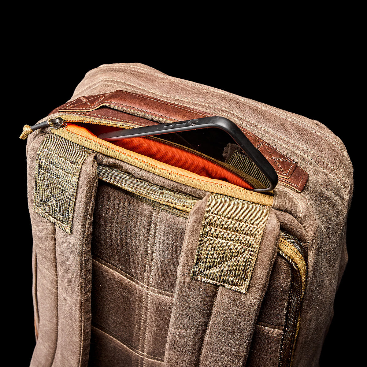 A rugged brown rucksack with a quilted design, viewed from the top angle. One compartment is slightly open, showing an orange interior and the edge of a tablet. Made from waxed canvas, this GR1 USA - Heritage by GORUCK boasts padded shoulder straps for comfort.