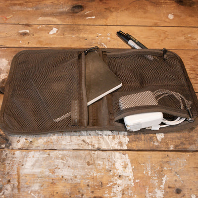 The GORUCK Wire Dopp, crafted from waxed canvas, is spread open on a rustic wooden surface. Inside, it reveals a notebook, charger, and pen, while additional compartments are designed to store all your essential travel items. Nearby on the table rest two pens.