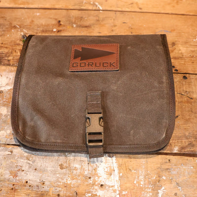 The Wire Dopp - Waxed Canvas by GORUCK, featuring a leather logo and buckle closure, rests on a wooden surface, perfect for organizing your travel essentials.