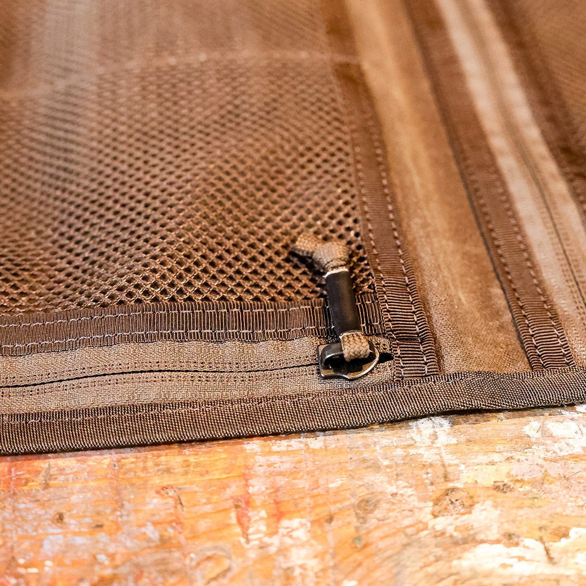 Close-up of the Wire Dopp - Waxed Canvas by GORUCK, featuring a brown mesh fabric with a zipper and toggle fastening on a wooden surface, highlighting its intricate stitching. Perfect for travel essentials, the textured design ensures optimal organization while providing durability similar to waxed canvas.