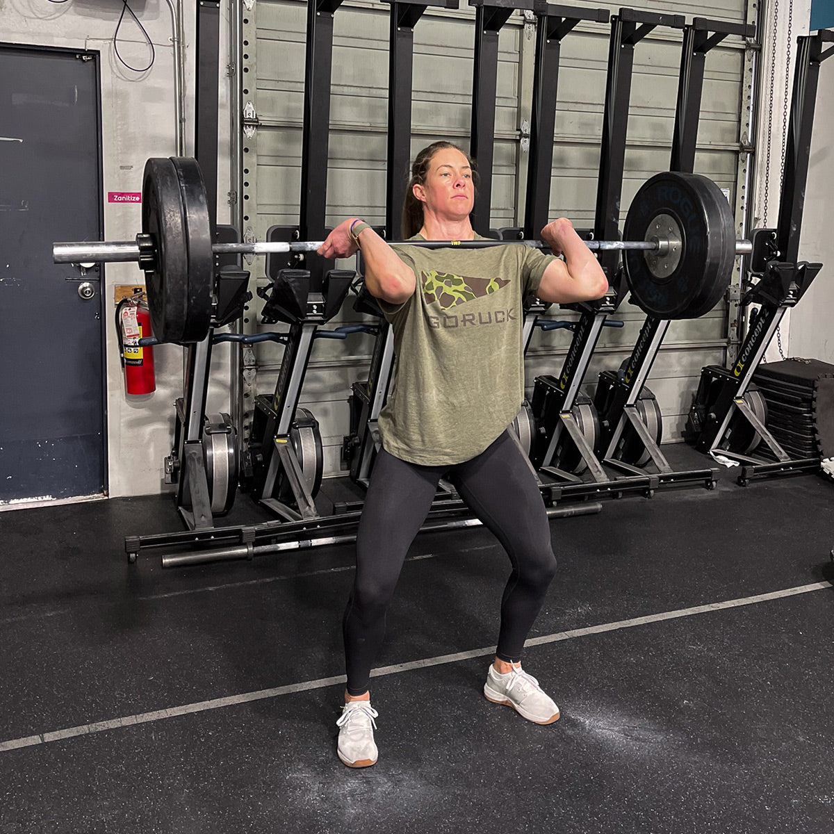 In the gym, someone is executing a front squat with a heavy barbell, channeling strength rooted in Special Forces inspiration. They are wearing the GORUCK Spearhead Tee - Tri-Blend in green and black leggings, positioned in front of stationary bikes on the concrete floor surrounded by metal equipment.