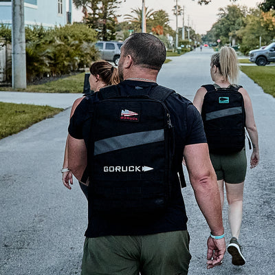 Three people with backpacks and Men’s Performance Tee - ToughMesh shirts stroll down a suburban street lined with trees and houses, their attire as durable as if they were trekking through the Italian Alps.