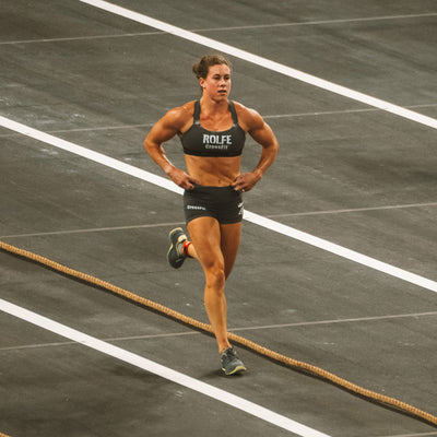 A person running on an indoor track, dressed in athletic shorts and GORUCK's Power Bra - Toughflex designed for high-impact movements. The track's surface features white lane markings and a coiled rope on one side, set against a grayish-brown background, ensuring comfort with its ToughStretch Fabric.