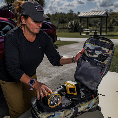 At a picnic area, a person is carefully placing equipment into the Bullet Ruck Classic - Cordura daypack by GORUCK. The padded straps promise comfort, while cars and a shelter provide the backdrop to this outdoor scene.