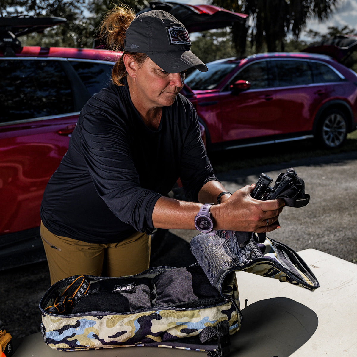 A person is packing camping gear into a Bullet Ruck Classic - Cordura by GORUCK, featuring padded straps, beside a red car in an outdoor setting.