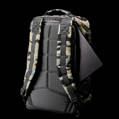 The GR1 USA - Cordura (The Original Ruck) by GORUCK is a camouflage backpack featuring padded straps, expertly crafted with American manufacturing precision. A tablet subtly emerges from a side pocket, capturing the stealth of Special Forces against a black background.