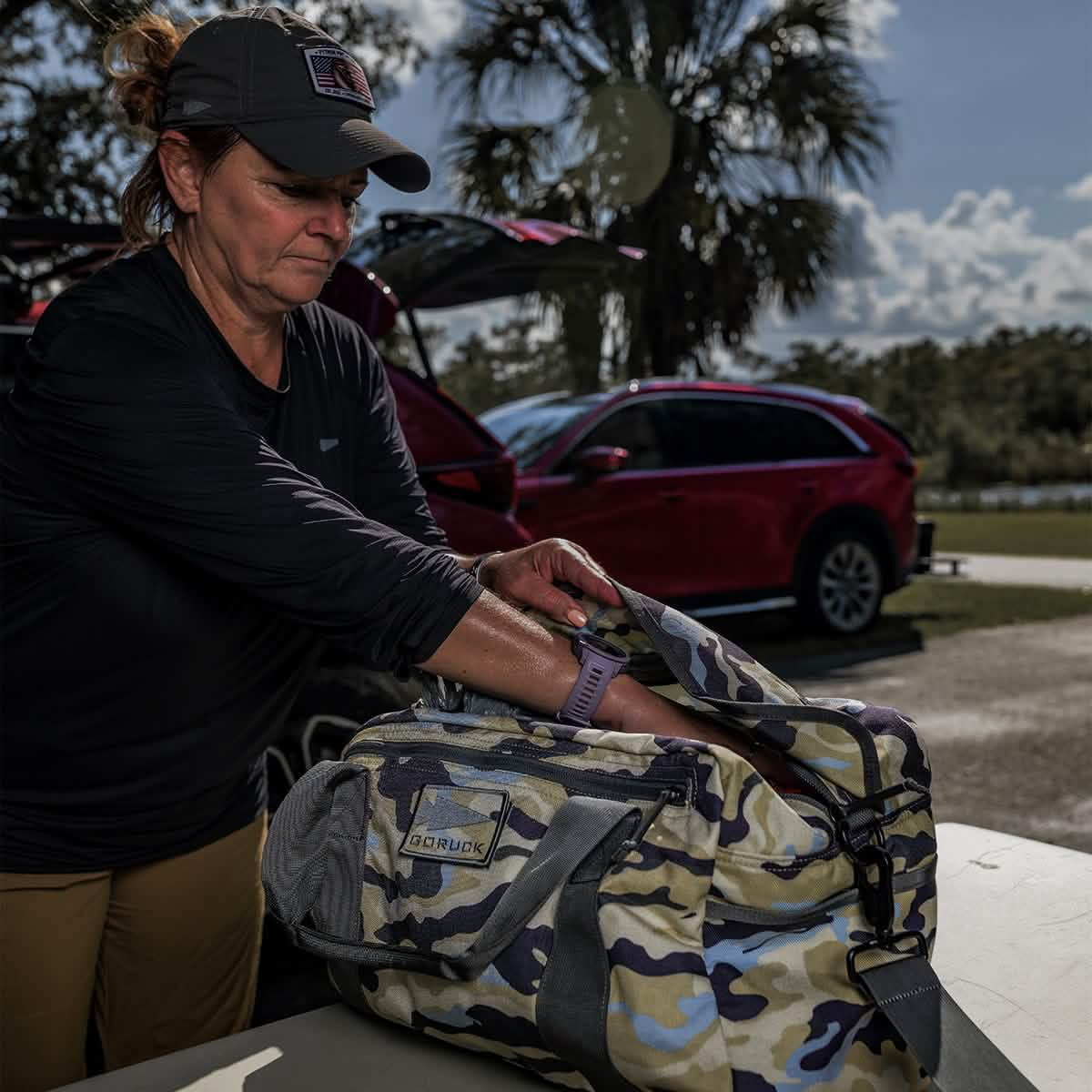 Someone is carefully packing a camo Kit Bag (Includes Shoulder Strap) on a table near a red car, outdoors, gearing up for an adventure.