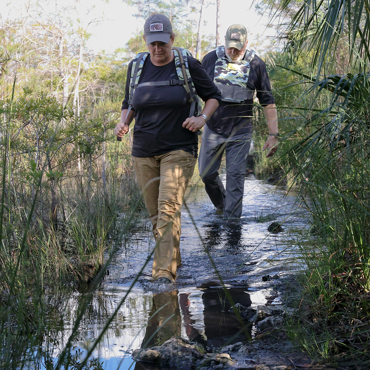 Two adventurers in outdoor gear navigate a muddy, forested trail surrounded by tall grass and trees while wearing comfortable GORUCK backpacks equipped with the Training Weight Vest 2.0. The scene evokes an adventurous trek in a natural environment.