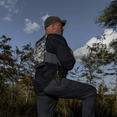 A man wearing outdoor gear, including a GORUCK Training Weight Vest 2.0 with comfortable shoulder straps and a hat, stands in a grassy field under a cloudy blue sky, suggesting a hiking or nature scene.