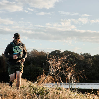 A man jogs outdoors near a body of water, wearing a cap and sports clothing. He carries a cup while showcasing the GORUCK Training Weight Vest 2.0, which features comfortable shoulder straps and a camouflage pattern. The background is adorned with trees beneath a mostly cloudy sky.