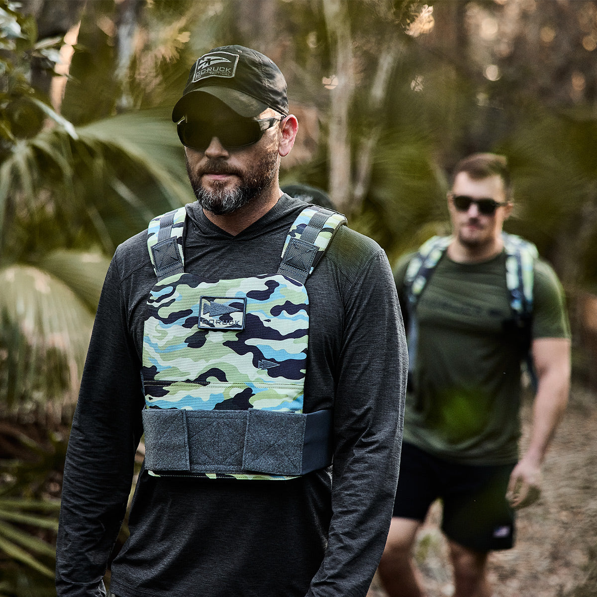 Two men walking through a forest trail. The man in the foreground is wearing the Training Weight Vest 2.0 by GORUCK, which features comfortable shoulder straps, along with sunglasses and a black cap, while the man behind is wearing a green shirt and backpack. Both appear to be hiking.