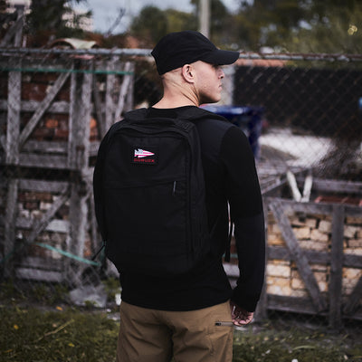 A man wearing a black backpack and cap stands by a fenced area with wooden pallets, dressed in the stylish Men’s Commando Long Sleeve - Merino Wool by GORUCK.