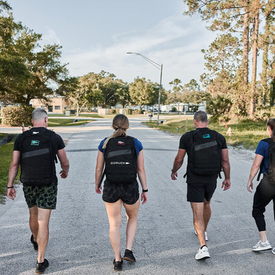 Four individuals stroll down a suburban street, each wearing backpacks adorned with the GORUCK Spearhead Patch. They are dressed in athletic wear and appear to be participating in a fitness activity. Trees and houses line the street under a clear blue sky.