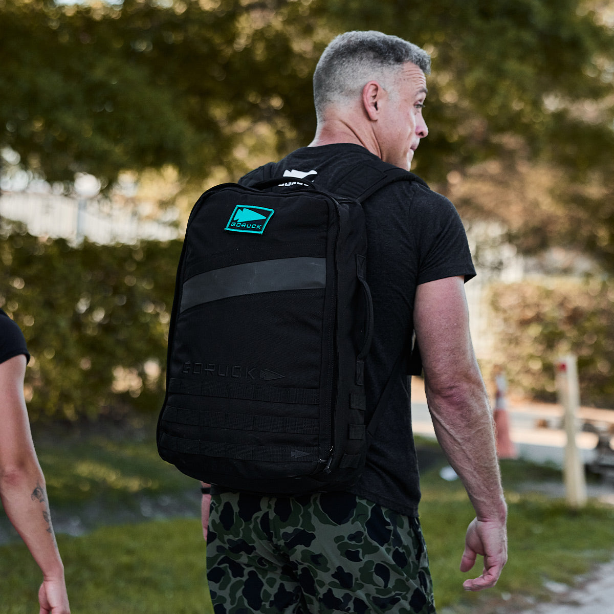 A man strides outdoors, adorned in a black t-shirt and camouflage pants, with a large black backpack that showcases the GORUCK Spearhead patch. The backdrop of lush greenery and a blurred fence adds an adventurous touch to his journey.