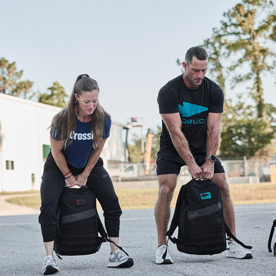 Two individuals are exercising outdoors, each lifting a weighted backpack with the Patch - GORUCK Spearhead by GORUCK. They stand on a concrete surface, framed by a white building and trees in the background, both focused intently on their workout.