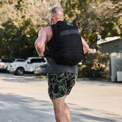 A jogger wearing a black backpack features an embroidered patch with the GORUCK Spearhead logo. They are styled in a sleeveless shirt and camouflage shorts, with trees and parked cars in the background enhancing the peaceful ambiance of their workout.