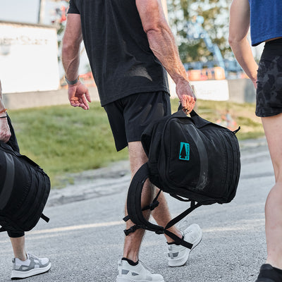 Three individuals strolling outdoors, each carrying a large black GORUCK backpack, adorned with an embroidered GORUCK Spearhead patch. They are dressed in casual athletic attire. The blurred urban background, interspersed with patches of grass, reflects their energetic lifestyle.