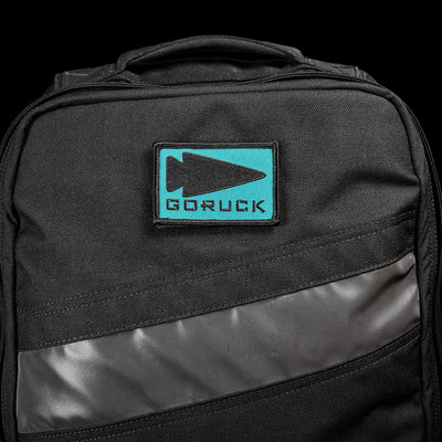 Close-up of a black backpack adorned with the Patch - GORUCK Spearhead, showcasing an embroidered logo patch with a blue background and black arrowhead. The backpack features a diagonal gray stripe detail and VELCRO backing, complete with a top handle, set against a black background.