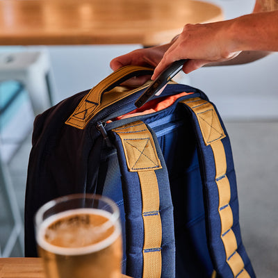 A person holding a GORUCK GR1 USA - Cordura backpack in navy and yellow, evocative of the durability of Special Forces gear, sits beside a glass of beer on a wooden table.