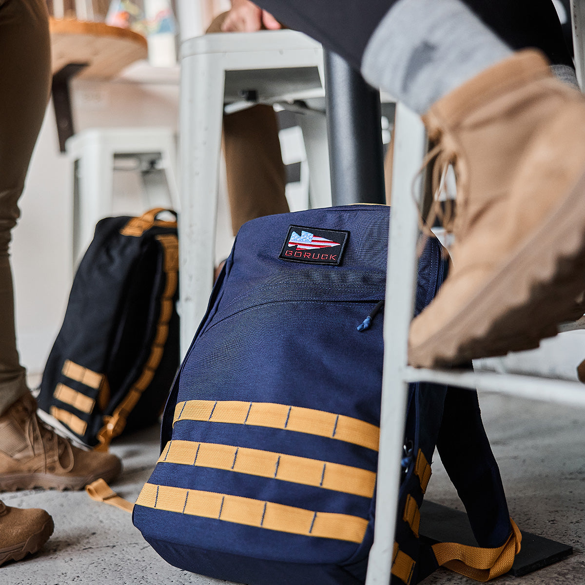 A GR1 USA - Cordura backpack in navy and tan from GORUCK rests on the floor next to people sitting on stools, their boots visible—ideal for those prepared to tackle the GORUCK Challenge.