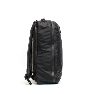 Side view of the stylish, black GR1 USA - X-PAC rucksack from GORUCK, showcasing its textured fabric. It includes waterproof compartments, a top handle, and adjustable shoulder straps. The compact and modern design makes it perfect for everyday use or travel.