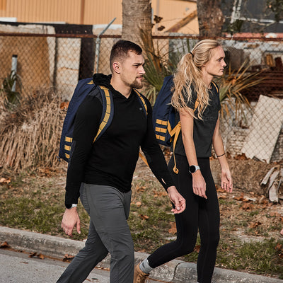 Two individuals stroll down the sidewalk, each carrying their GR1 USA - Cordura (The Original Ruck) by GORUCK, crafted with Special Forces-grade construction. Their rucksacks boast a bombproof laptop compartment, safeguarding their belongings as they move through the bustling street.