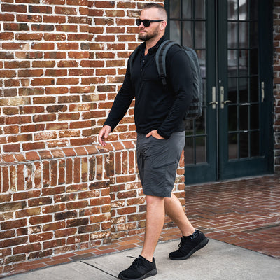 A man wearing sunglasses, a black long-sleeve shirt, and GORUCK’s Men’s Challenge Shorts - Lightweight ToughDry® walks on the sidewalk. He carries a backpack and is passing by a brick building with large windows and a door, his black shoes tapping lightly against the pavement.