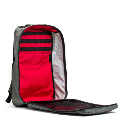 The GR1 USA - X-PAC rucksack by GORUCK stands upright with its front panel fully unzipped, revealing a red interior featuring several pockets and compartments for organization. Made from durable materials, this gray backpack is not only stylish but also waterproof, keeping your belongings dry.