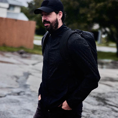 A man with a beard, dressed in a black cap and jacket, walks casually on a damp paved path. His hands tucked into his pockets, he carries a GR1 USA - X-PAC rucksack from GORUCK on his back. Trees and a wooden fence provide a backdrop beneath the cloudy sky.