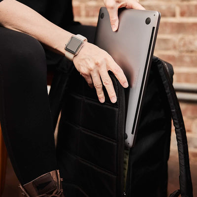 A person wearing a smartwatch is placing a laptop into the GR1 USA - X-PAC rucksack by GORUCK, which is made from waterproof X-PAC material. The setting appears indoors with a brick wall in the background. They are seated, wearing dark clothing and brown shoes.