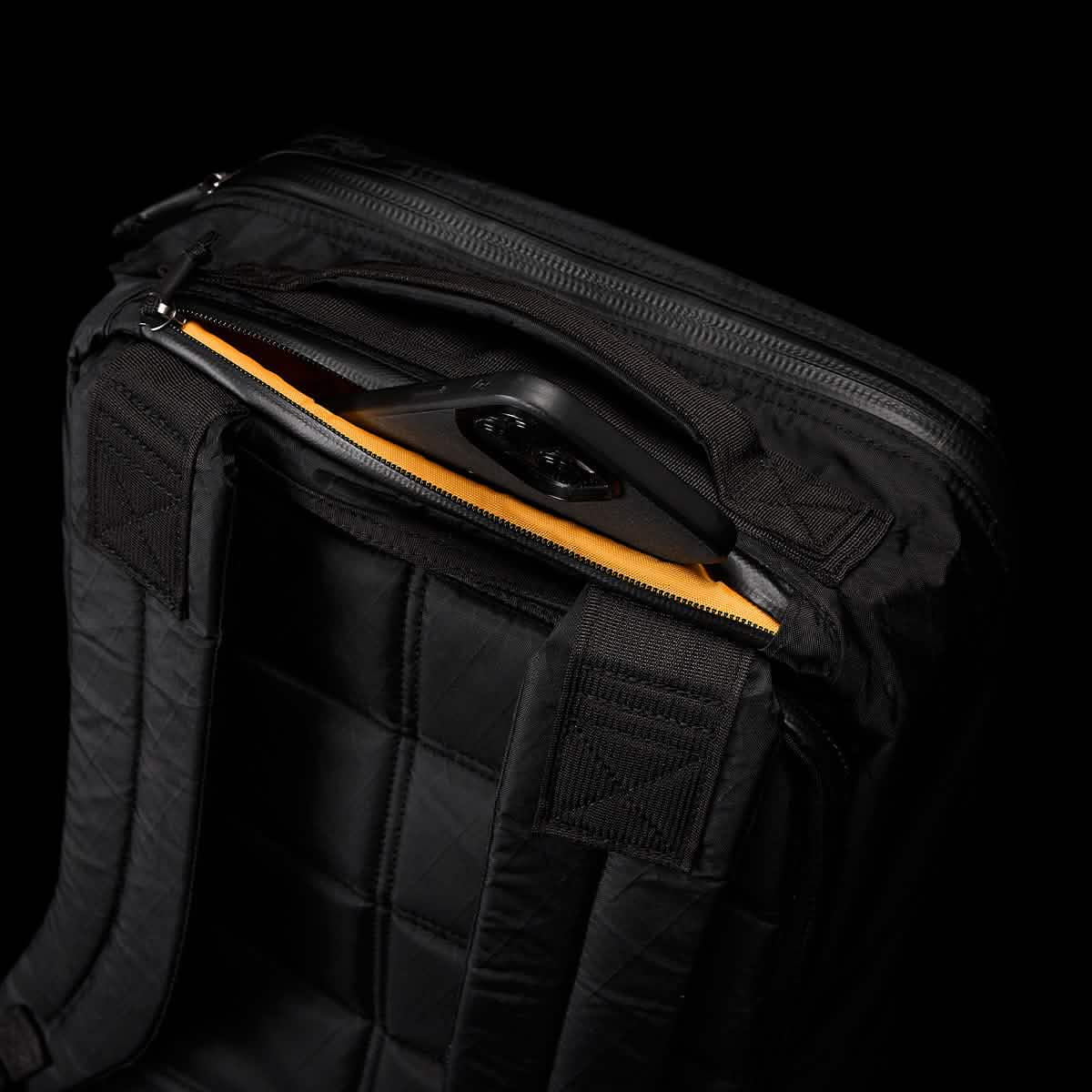 In a dimly lit setting, the GR1 USA - X-PAC rucksack by GORUCK is displayed. The unzipped top pocket reveals an orange interior with a black phone case partially visible inside. The waterproof backpack, made from X-PAC material, features its straps prominently in the foreground.