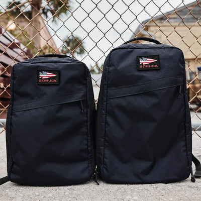 Two GR1 USA - X-PAC rucksacks, each featuring GORUCK patches, stand upright against a chain-link fence. Crafted from durable X-PAC material, these black backpacks are waterproof and ideal for urban adventures. The background suggests a cityscape with palm trees and buildings partially visible.