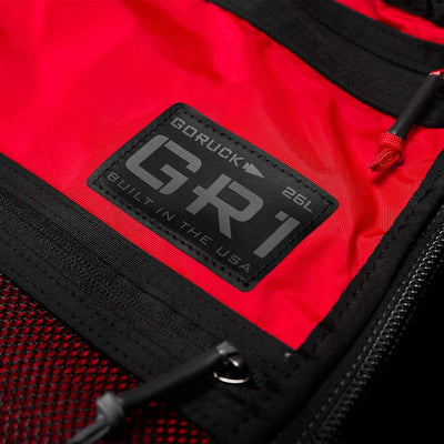 Close-up of a red GORUCK GR1 USA - X-PAC rucksack showcasing a black label with "GR1 26L Built in the USA" text. The label is sewn onto the waterproof X-PAC fabric and is accented by sleek black zippers and mesh pocket detailing.