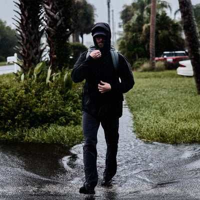 A person strolls along a wet street during rainfall, clad in a black raincoat with a hood and carrying the GR1 USA - X-PAC rucksack by GORUCK. The background showcases palm trees and parked cars.