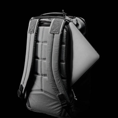 The GORUCK GR1 USA - X-PAC, a sleek gray rucksack, is displayed against a black background. It features waterproof X-PAC material and padded shoulder straps. A laptop is partially visible as it is slid into the secure back compartment of this backpack with its textured, quilted design.