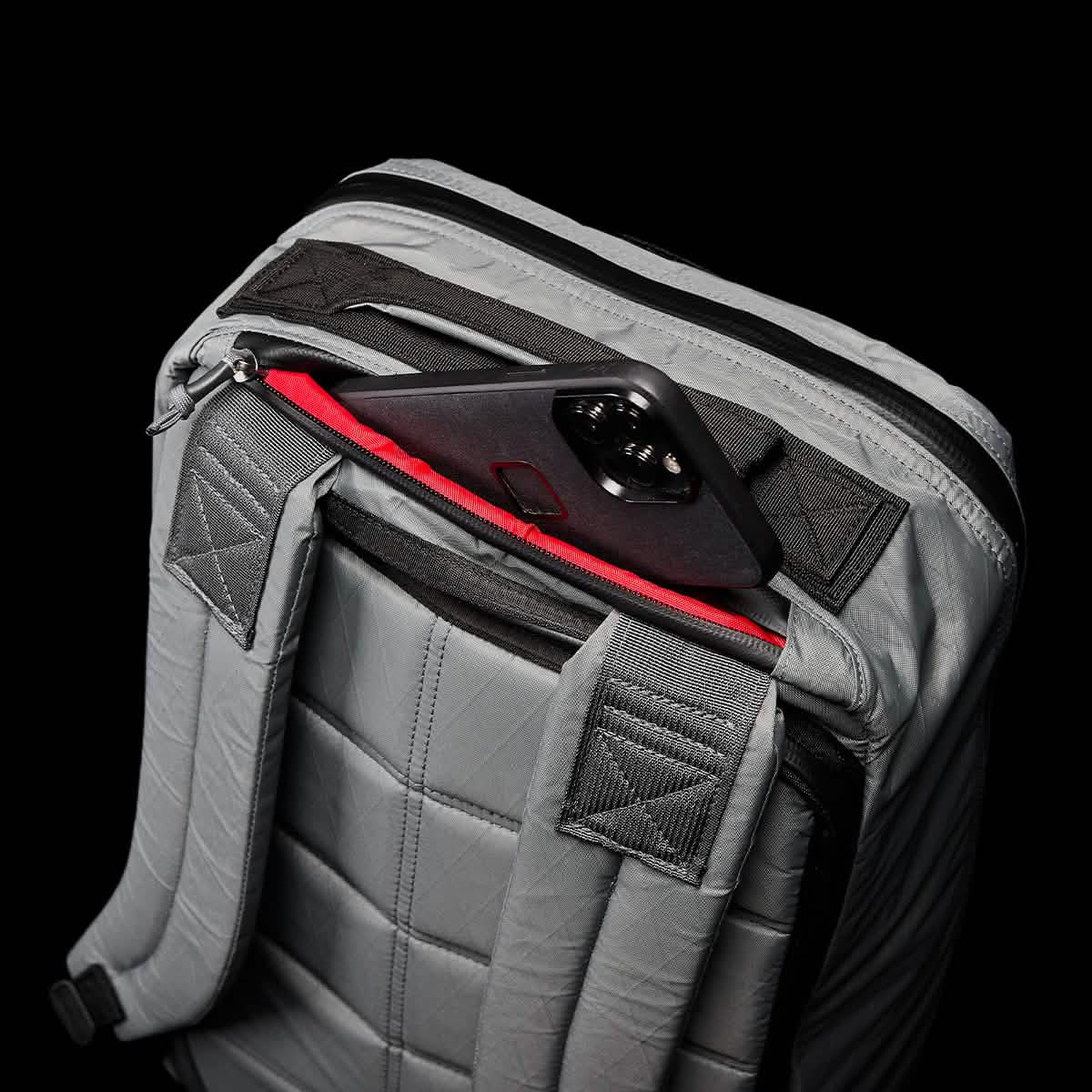 The GORUCK GR1 USA - X-PAC rucksack, featuring quilted straps and a sleek, waterproof design, is partially opened to reveal a black smartphone inside the red-lined pocket, set against a black background.