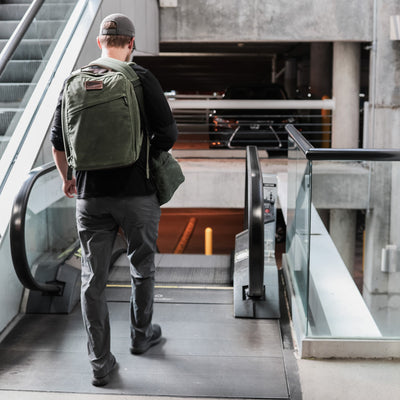 A man wearing a cap, black shirt, and gray pants stands on a moving walkway with a green GR1 USA - Heritage backpack and duffel bag from GORUCK. He is inside a concrete parking area with escalators and parked cars visible in the background.