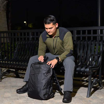 A person wearing a green and black shirt sits on a bench at night, looking down as they reach into their GR1 USA - Dyneema rucksack by GORUCK. The scene is dimly lit, with a few lights illuminating the background.